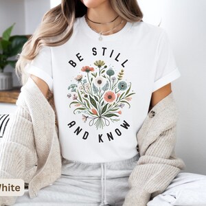 May include: White t-shirt with a floral design and the text "Be Still and Know" in black.
