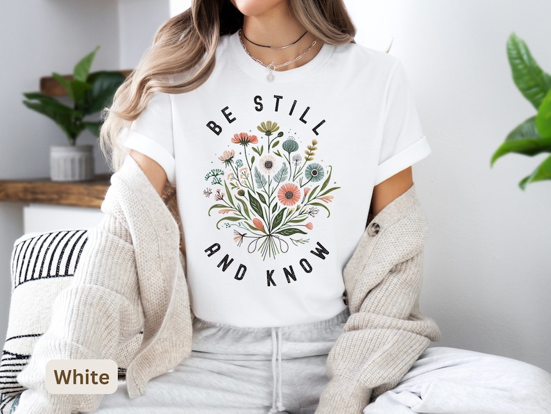 May include: White t-shirt with a floral design and the text "Be Still and Know" in black.