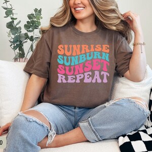Op de afbeelding: Espresso-kleurig t-shirt met de tekst "Sunrise Sunburn Sunset Repeat" in een retro lettertype. De tekst is oranje, turquoise, roze en paars. Het shirt wordt gedragen met een blauwe jeans.