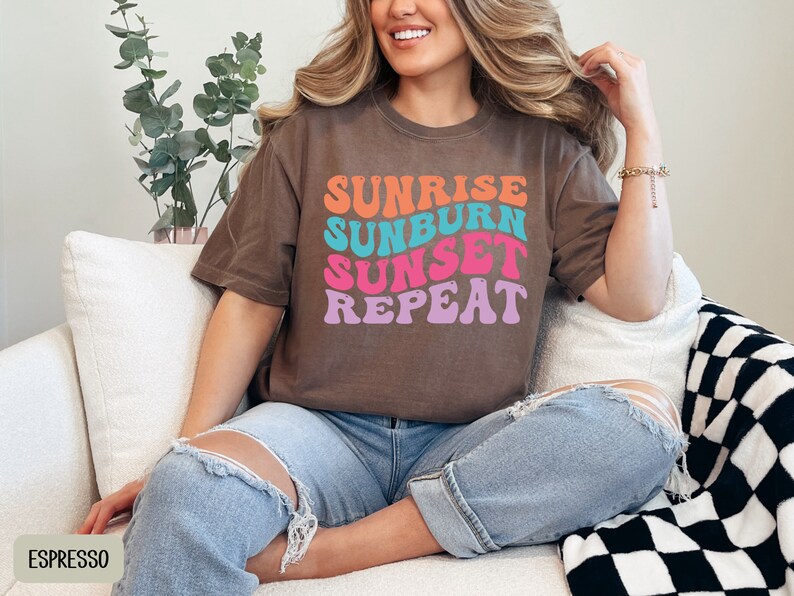 Op de afbeelding: Espresso-kleurig t-shirt met de tekst "Sunrise Sunburn Sunset Repeat" in een retro lettertype. De tekst is oranje, turquoise, roze en paars. Het shirt wordt gedragen met een blauwe jeans.