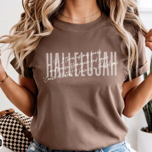 May include: A brown short-sleeve t-shirt featuring the word HALLELUJAH in large white block letters, with the cursive text 'So I throw up my hands and praise You again and again 'Cause all that I have is a hallelujah' layered over it.