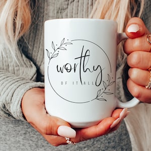 Puede incluir: Taza de cerámica blanca con texto negro y diseño floral que dice "worthy of it all".
