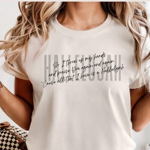 May include: An ivory short-sleeved t-shirt featuring the word HALLELUJAH in large gray capital letters. Overlaid in black cursive script is the text: So I throw up my hands and praise You again and again 'Cause all that I have is a Hallelujah.