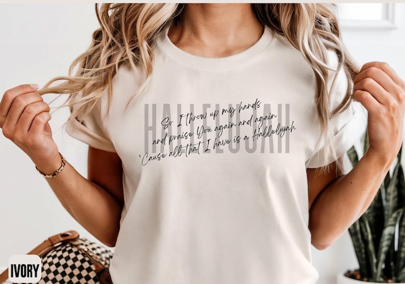 May include: An ivory short-sleeved t-shirt featuring the word HALLELUJAH in large gray capital letters. Overlaid in black cursive script is the text: So I throw up my hands and praise You again and again 'Cause all that I have is a Hallelujah.
