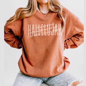 May include: A rust-colored crewneck sweatshirt featuring the word HALLELUJAH in large white block letters, with smaller cursive white text layered over the center that reads So I throw up my hands and praise Him again and again 'Cause all that I have is a hallelujah.