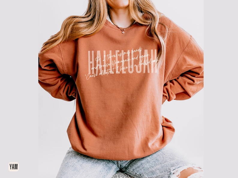 May include: A rust-colored crewneck sweatshirt featuring the word HALLELUJAH in large white block letters, with smaller cursive white text layered over the center that reads So I throw up my hands and praise Him again and again 'Cause all that I have is a hallelujah.