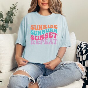Op de afbeelding: Lichtblauw T-shirt met de tekst "Sunrise Sunburn Sunset Repeat" in oranje, turquoise, roze en paars. Het T-shirt wordt gedragen met een lichtblauwe jeans. Het thema is zomer en vakantie.