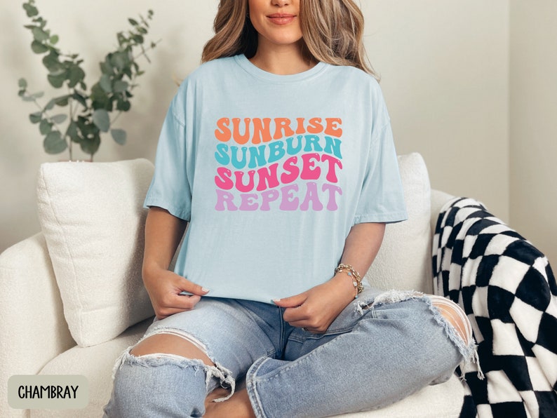 Op de afbeelding: Lichtblauw T-shirt met de tekst "Sunrise Sunburn Sunset Repeat" in oranje, turquoise, roze en paars. Het T-shirt wordt gedragen met een lichtblauwe jeans. Het thema is zomer en vakantie.