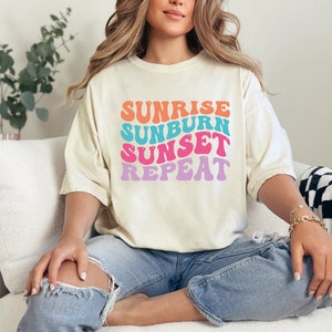 Op de afbeelding: Cr&egrave;mekleurig T-shirt met de tekst "Sunrise Sunburn Sunset Repeat" in een retro lettertype. De tekst is oranje, blauw, roze en paars. Het shirt wordt gecombineerd met een blauwe jeans. Het woord "IVORY" staat linksonder.