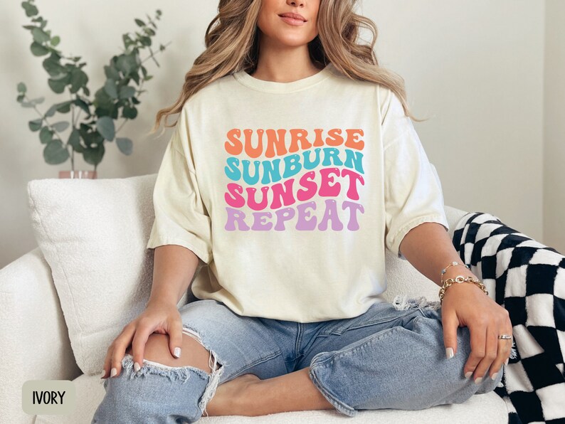 Op de afbeelding: Cr&egrave;mekleurig T-shirt met de tekst "Sunrise Sunburn Sunset Repeat" in een retro lettertype. De tekst is oranje, blauw, roze en paars. Het shirt wordt gecombineerd met een blauwe jeans. Het woord "IVORY" staat linksonder.