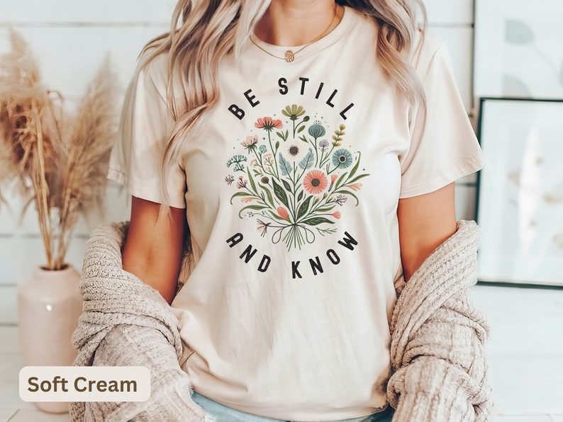 May include: A soft cream colored t-shirt with a floral design and the text "Be Still and Know" printed on the front.