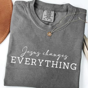 May include: A gray t-shirt with the words "Jesus changes EVERYTHING" in white script. A gold pendant necklace and a dried palm leaf are also visible. The shirt is a Comfort Colors brand, size S-P-CH.