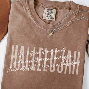 May include: A brown short-sleeved t-shirt with a crew neckline featuring the word HALLELUJAH in large light-colored block letters and white cursive text that reads: So I throw up my hands and praise You again and again 'Cause all that I have is a Hallelujah.