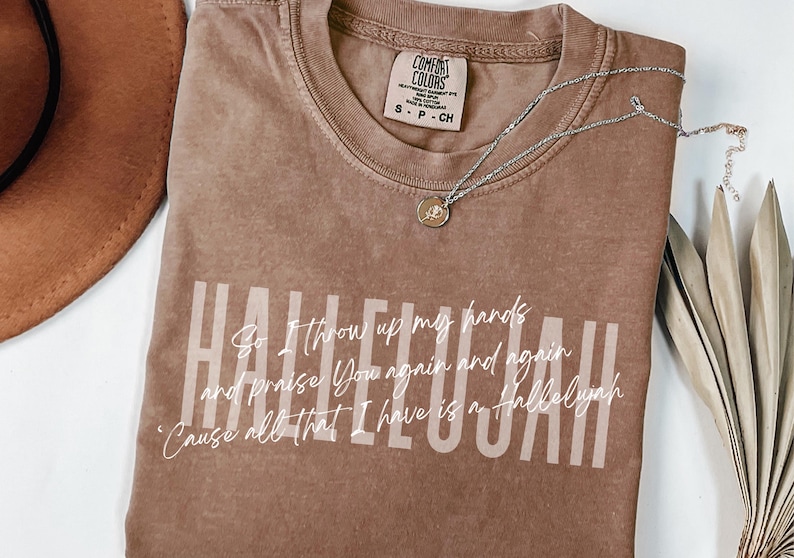 May include: A brown short-sleeved t-shirt with a crew neckline featuring the word HALLELUJAH in large light-colored block letters and white cursive text that reads: So I throw up my hands and praise You again and again 'Cause all that I have is a Hallelujah.
