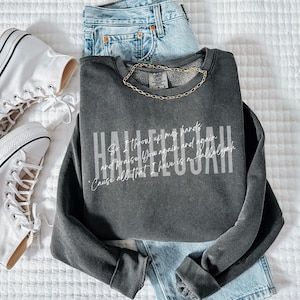 May include: A charcoal gray crewneck sweatshirt featuring the white text Hallelujah and smaller script lyrics. The garment is paired with light blue denim jeans and white high-top canvas sneakers with black trim and white laces.