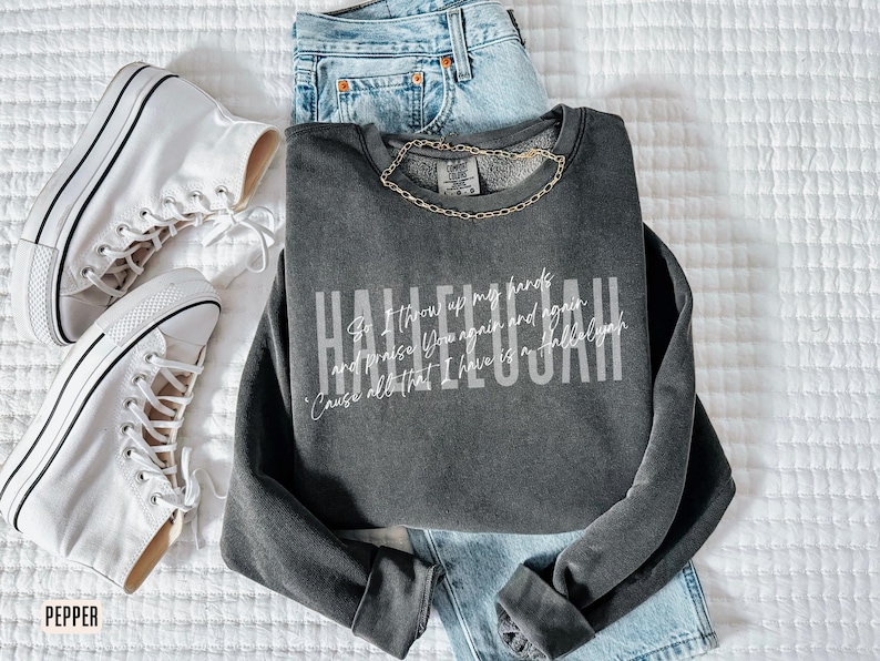 May include: A charcoal gray crewneck sweatshirt featuring the white text Hallelujah and smaller script lyrics. The garment is paired with light blue denim jeans and white high-top canvas sneakers with black trim and white laces.