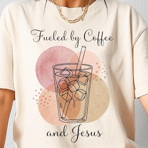 Puede incluir: Camiseta color crema con la frase "Fueled by Coffee and Jesus" en tipografía cursiva. El diseño incluye un dibujo lineal de un vaso de café helado con cubitos de hielo y una pajita, sobre círculos de acuarela.