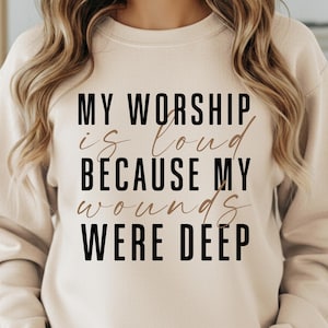 May include: Sand-colored sweatshirt with the text "MY WORSHIP is loud BECAUSE MY wounds WERE DEEP" in black and brown lettering. The sweatshirt is a neutral color and has a crew neck.