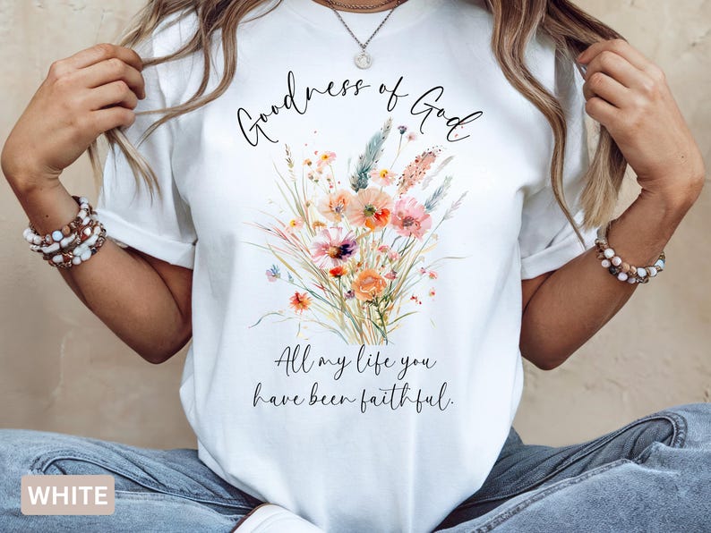 May include: White t-shirt featuring a watercolor floral design in shades of orange, pink, and green. The shirt has the text "Goodness of God" above the floral arrangement, and "All my life you have been faithful" below.