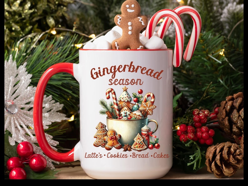 May include: A white ceramic mug with a red interior and handle, featuring the text "Gingerbread season" and gingerbread-themed illustrations. The mug is filled with marshmallows, a gingerbread man, and candy canes. The mug is surrounded by Christmas decorations.