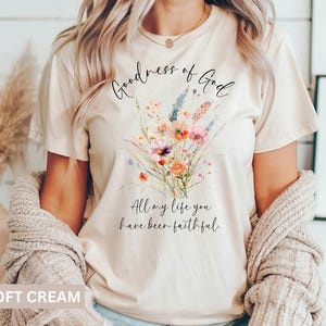 May include: Soft cream-colored t-shirt with a floral bouquet design. The bouquet features orange, pink, and purple flowers with green stems. The text "Goodness of God" and "All my life you have been faithful" are written in black script.