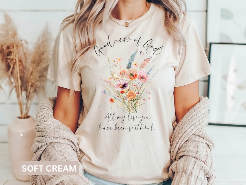 May include: Soft cream-colored t-shirt with a floral bouquet design. The bouquet features orange, pink, and purple flowers with green stems. The text "Goodness of God" and "All my life you have been faithful" are written in black script.