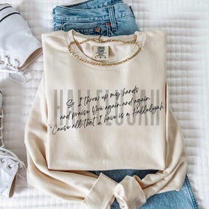 May include: A cream-colored crewneck sweatshirt featuring black cursive text that reads, So I throw up my hands and praise You again and again 'Cause all that I have is a Hallelujah, layered over faint gray text reading HALLELUJAH.