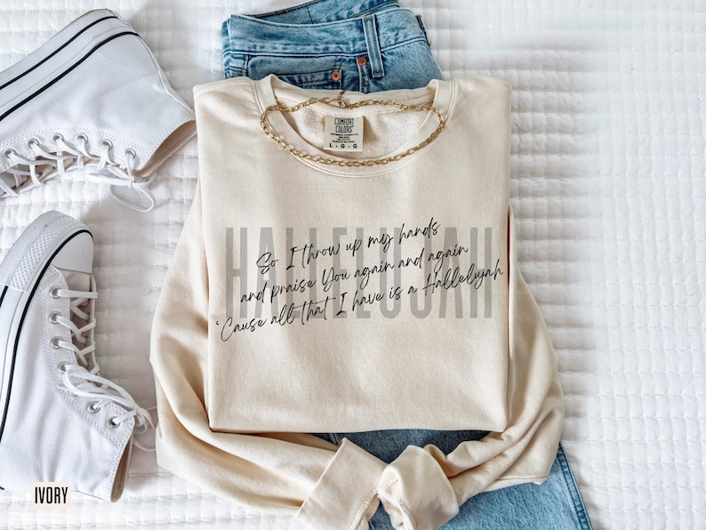 May include: A cream-colored crewneck sweatshirt featuring black cursive text that reads, So I throw up my hands and praise You again and again 'Cause all that I have is a Hallelujah, layered over faint gray text reading HALLELUJAH.