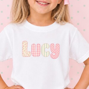 以下が含まれることがあります： 大きな丸みを帯びた文字で「Lucy」とプリントされた白い半袖Tシャツ。各文字には異なるパターンが施されており、Lはピンクの花柄、uはピンクのギンガムチェック、cは緑のストライプ、yはピンクの水玉模様になっています。