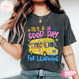 Op de afbeelding: Een antracietgrijs t-shirt met een afbeelding van een gele schoolbus en de tekst It's A GOOD DAY FOR LEARNING. De bus bevat illustraties van kinderen en een leraar, versierd met kleurrijke sterren, planeten en ruimtevaartmotieven.