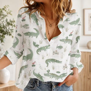 May include: A white long-sleeve button-down shirt featuring an all-over print of light green crocodiles, small pink birds, and green reeds. The shirt has a classic collar, a front button placket, and long sleeves with cuffs that are rolled up.