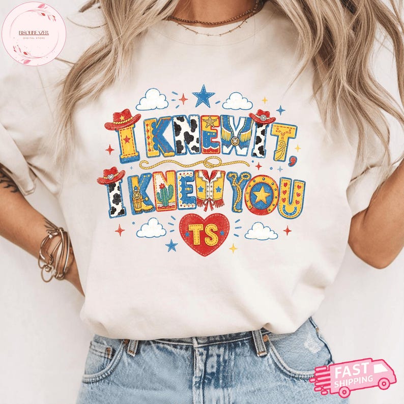 May include: A cream-colored t-shirt featuring a colorful graphic with the text I KNEW IT, I KNEW YOU and TS inside a red heart. The design includes cowboy hats, a cactus, stars, clouds, and rope motifs in shades of red, blue, yellow, and white.