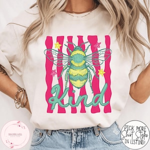 Puede incluir: Camiseta color crema con un diseño de abeja colorida y la palabra "Kind" en una fuente cursiva. La abeja es amarilla y verde, sobre un fondo de rayas rosas. La camiseta tiene cuello redondo.