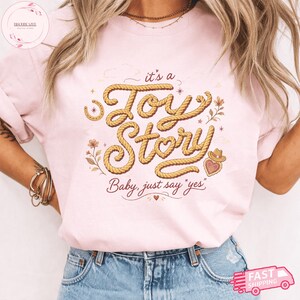 May include: A light pink t-shirt featuring a graphic design with the text "it's a Toy Story" written in a rope-like font, accompanied by the phrase "Baby, just say 'yes'" below. The design includes illustrations of a horseshoe, flowers, and a cowboy hat.