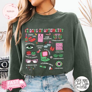 May include: Green sweatshirt featuring the phrase "12 Days of Optometry" and optometry-themed illustrations. The design includes eye charts, contact lenses, and eye examination tools. The sweatshirt is a comfortable, casual style.