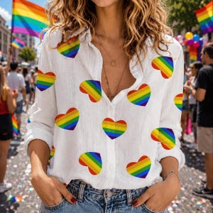 May include: White button-down shirt with long sleeves, adorned with rainbow heart patterns. The shirt is unbuttoned at the top, revealing a necklace. The background shows a street scene with a rainbow flag and people.