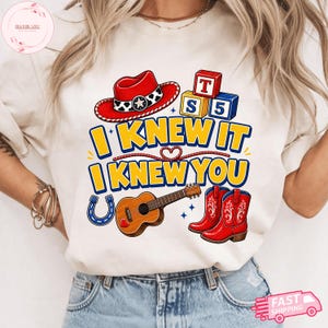 May include: A cream-coloured t-shirt featuring a graphic print with a red cowboy hat, alphabet blocks, a brown acoustic guitar, a blue horseshoe, and red cowboy boots. The text reads I KNEW IT I KNEW YOU in bold yellow letters with blue outlines.