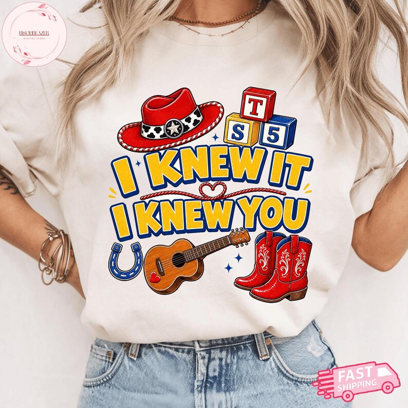 May include: A cream-coloured t-shirt featuring a graphic print with a red cowboy hat, alphabet blocks, a brown acoustic guitar, a blue horseshoe, and red cowboy boots. The text reads I KNEW IT I KNEW YOU in bold yellow letters with blue outlines.