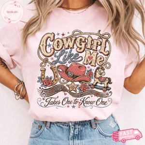 Op de afbeelding: Een lichtroze t-shirt met een grafisch ontwerp met de tekst "Cowgirl Like Me" en "Takes One to Know One". De illustratie bevat een lasso, een roze cowboyhoed, een hoefijzer, een hobbelpaard, muzieknoten en sterren.