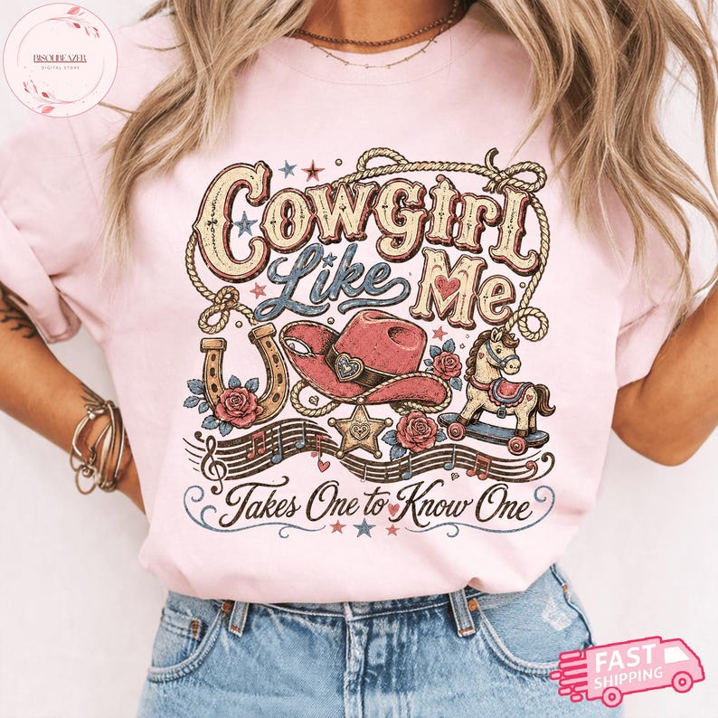 Op de afbeelding: Een lichtroze t-shirt met een grafisch ontwerp met de tekst "Cowgirl Like Me" en "Takes One to Know One". De illustratie bevat een lasso, een roze cowboyhoed, een hoefijzer, een hobbelpaard, muzieknoten en sterren.