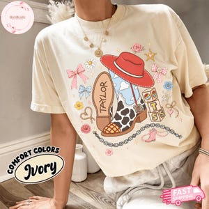 May include: An ivory short-sleeve t-shirt featuring a graphic print of a red cowboy hat, a cow-print cowboy boot with the name TAYLOR, and alphabet blocks with the letters T, S, and S. The design includes pink bows, flowers, and stars.
