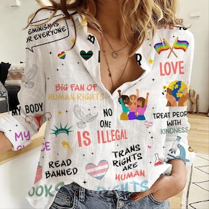 May include: White button-down shirt with multiple graphics and text. The shirt displays phrases such as "Feminism is for Everyone," "Big Fan of Human Rights," and "Trans Rights are Human." It also includes images of doves, hearts, and rainbow flags.