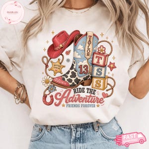 May include: A cream-colored t-shirt featuring a graphic print of a pink cowboy hat, a blue and white cow-print cowboy boot with the number 13, a sheriff badge labeled Besties Forever, three blocks labeled T S S, and the text Ride The Adventure Friends Forever.