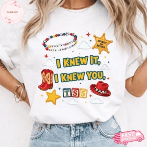May include: A white t-shirt featuring a graphic print with the text I KNEW IT, I KNEW YOU, TS5, INFINITY AND BEYOND, and YOU'VE GOT A FRIEND IN ME. The design includes a beaded bracelet, a sheriff star, red cowboy boots, a red hat, and blocks.