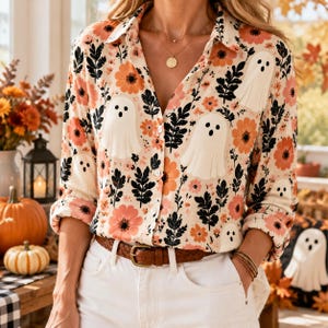 May include: A long-sleeved button-down shirt featuring a cream background with a repeating pattern of white ghosts, orange flowers, and black foliage. The shirt has a classic collar, a front button placket, and is tucked into white pants.