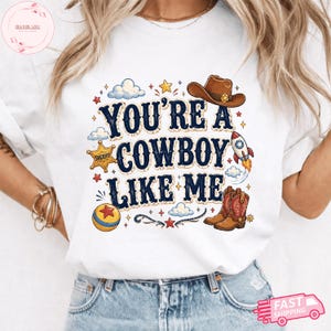 May include: A white t-shirt featuring the text "YOU'RE A COWBOY LIKE ME" in dark blue lettering. The design includes a brown cowboy hat, a yellow sheriff star badge, a red and white rocket, a pair of brown cowboy boots, and a colourful ball.