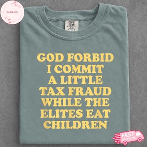 May include: A sage green crew neck t-shirt featuring the yellow text "GOD FORBID I COMMIT A LITTLE TAX FRAUD WHILE THE ELITES EAT CHILDREN" printed in a bold, serif font across the chest. The shirt includes a Comfort Colors brand label.