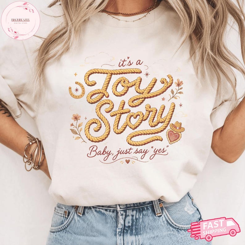 May include: A cream-colored t-shirt featuring a graphic design with the text "it's a Toy Story" written in a yellow rope-style font. The design includes small illustrations of a horseshoe, flowers, a heart with a cowboy hat, and the text "Baby, just say yes".