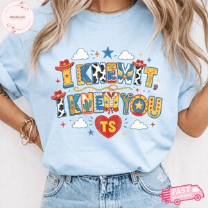 May include: A light blue t-shirt featuring a graphic print with the text I KNEW IT, I KNEW YOU and TS inside a red heart. The design includes cowboy hats, cow print patterns, a cactus, stars, clouds, and a lasso in red, yellow, and blue colors.
