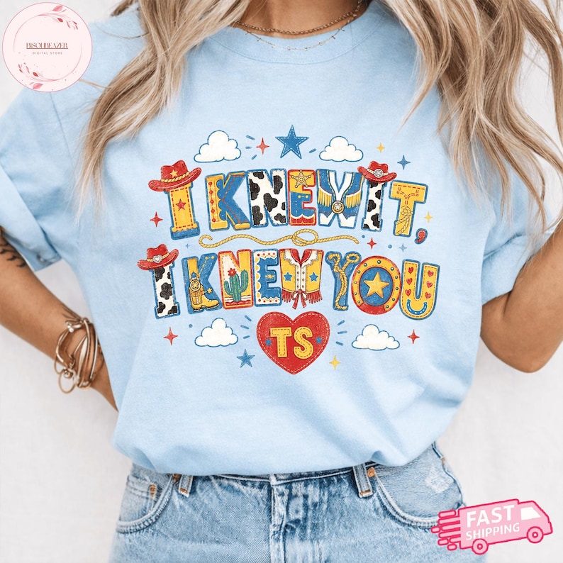 May include: A light blue t-shirt featuring a graphic print with the text I KNEW IT, I KNEW YOU and TS inside a red heart. The design includes cowboy hats, cow print patterns, a cactus, stars, clouds, and a lasso in red, yellow, and blue colors.